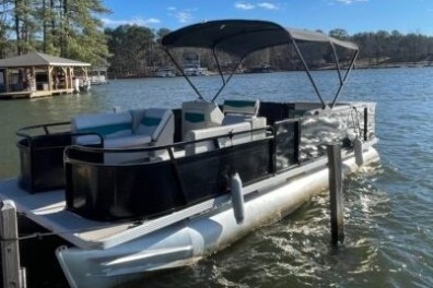 Pontoon boat with canopy docked on a lake near trees and cabin.