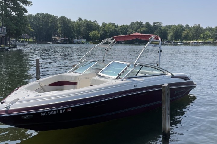 A white and maroon speedboat docked on a lake with trees in the background.