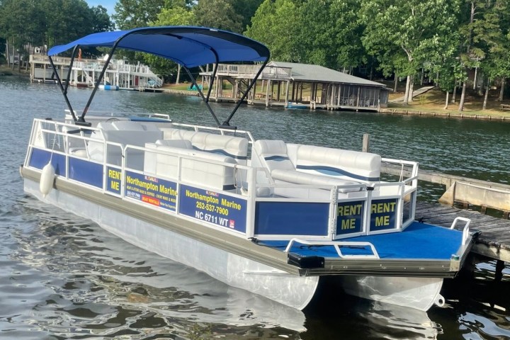 Blue pontoon boat with canopy docked by a lake with trees and houses in the background.
