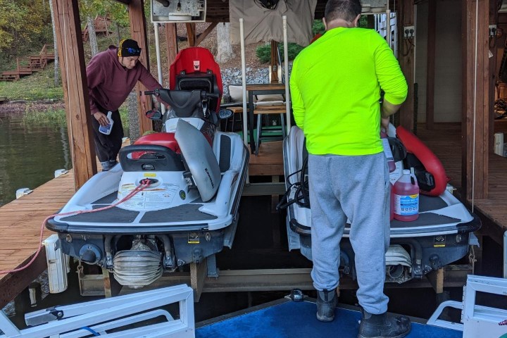 Two people working on jet skis in a covered dock area with various tools around.