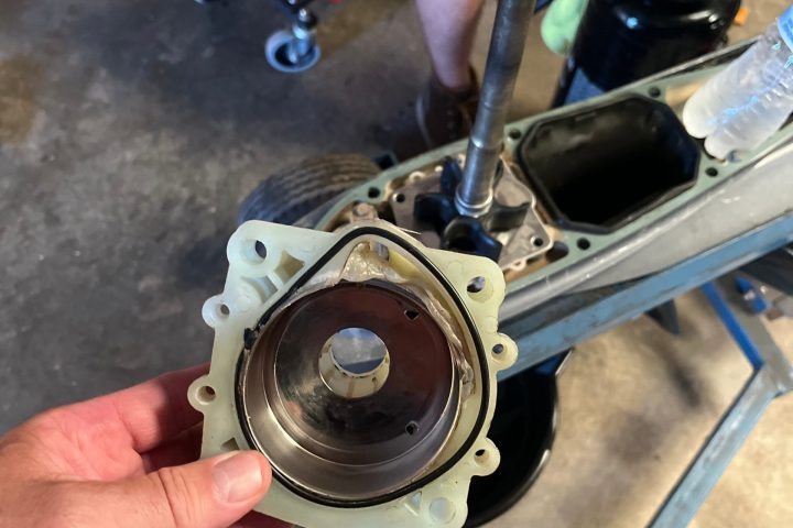 Person holding a mechanical part in a garage with tools and a seated person in the background.