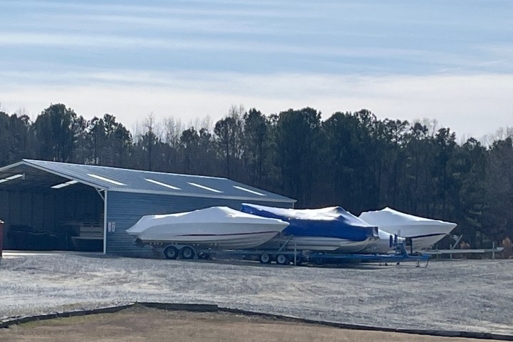 Three boats covered and parked outside a metal shed near a forested area.