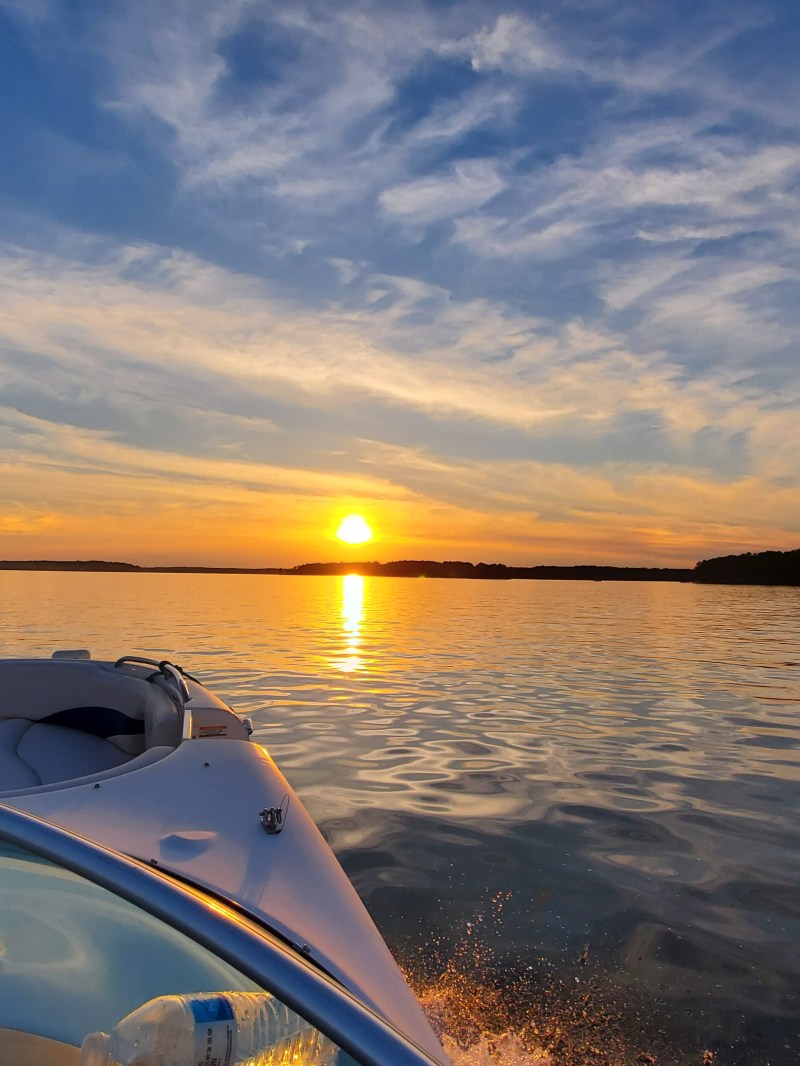 Boat on a lake at sunset with a colorful sky and trees on the horizon.