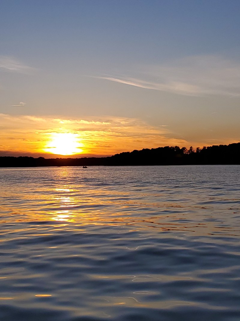 Sunset over a calm lake with distant tree line and colorful sky.